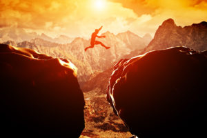 Man jumping over precipice between two rocky mountains at sunset. Freedom, risk, challenge, success.
